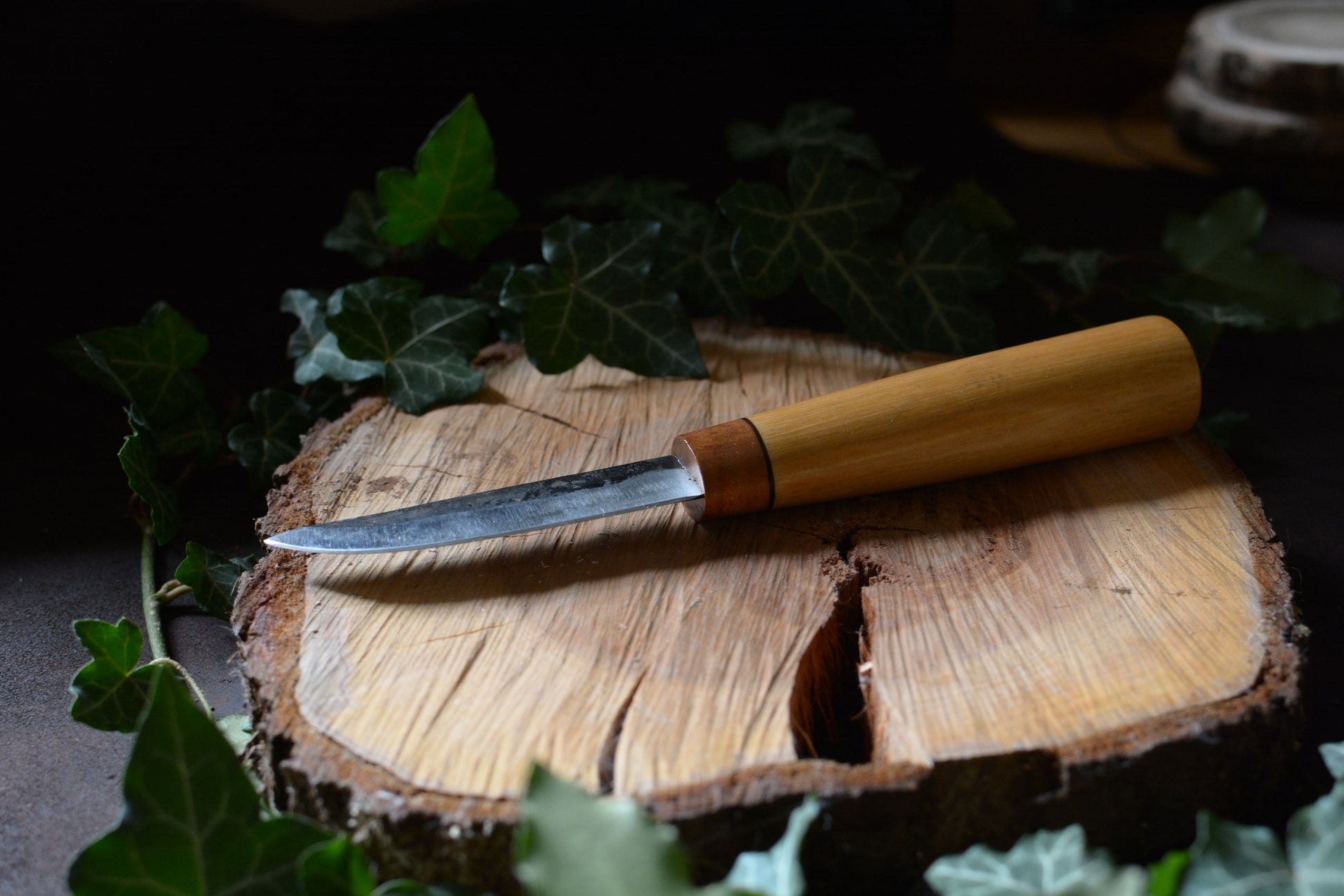 Vue sur l'épaisseur de la lame. Couteau forgé avec manche en charme et noyer posé sur un rondin en bois avec décor feuilles de lierre