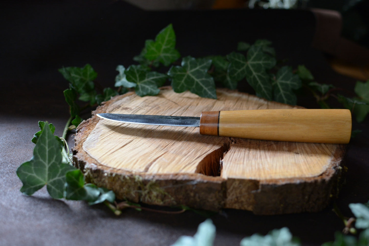Vue de l'épaisseur de lame. Couteau forgé avec manche en charme et noyer posé sur un rondin en bois avec décor feuilles de lierre