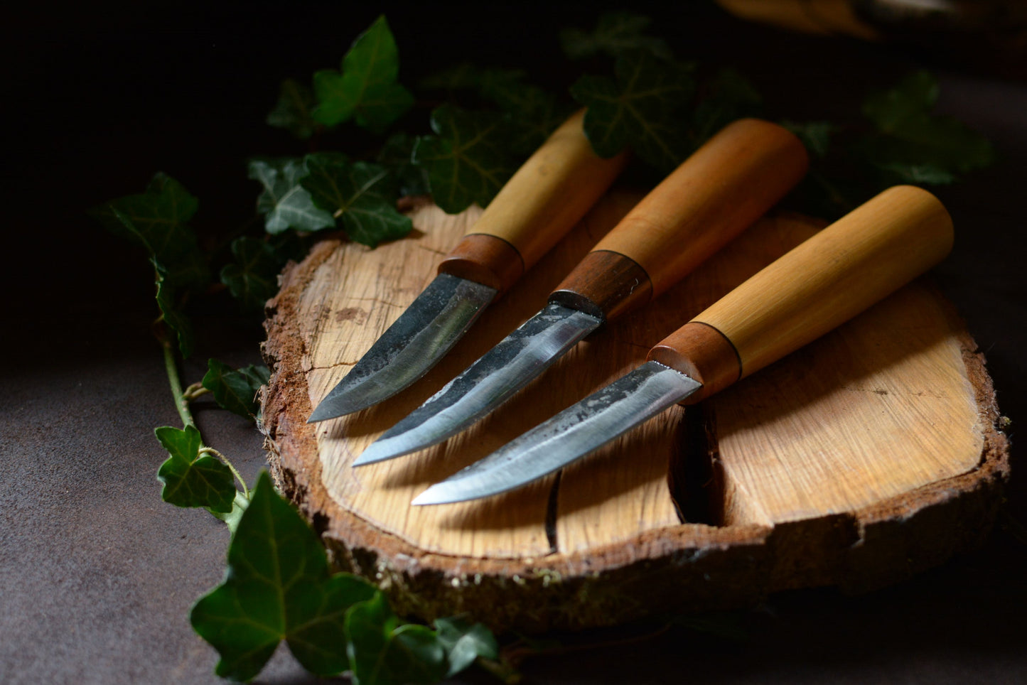Collection de 3 couteaux forgés avec manches en charme, érable et noyer posés sur un rondin en bois et décor en feuilles de lierre