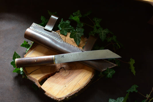 couteau artisanal scramasaxe en noyer et laiton et son étui en cuir posé sur un rondin en bois et feuilles de lierre